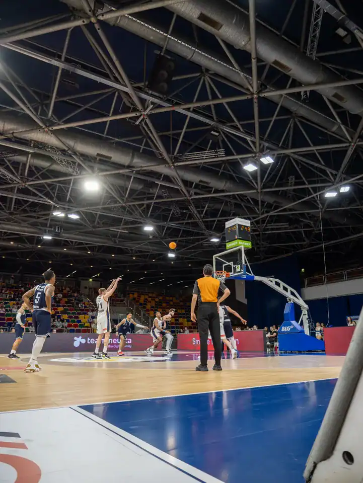 Blick vom Spielfeldrand auf Korb, Spieler und Dachkonstruktion der Basketballhalle.