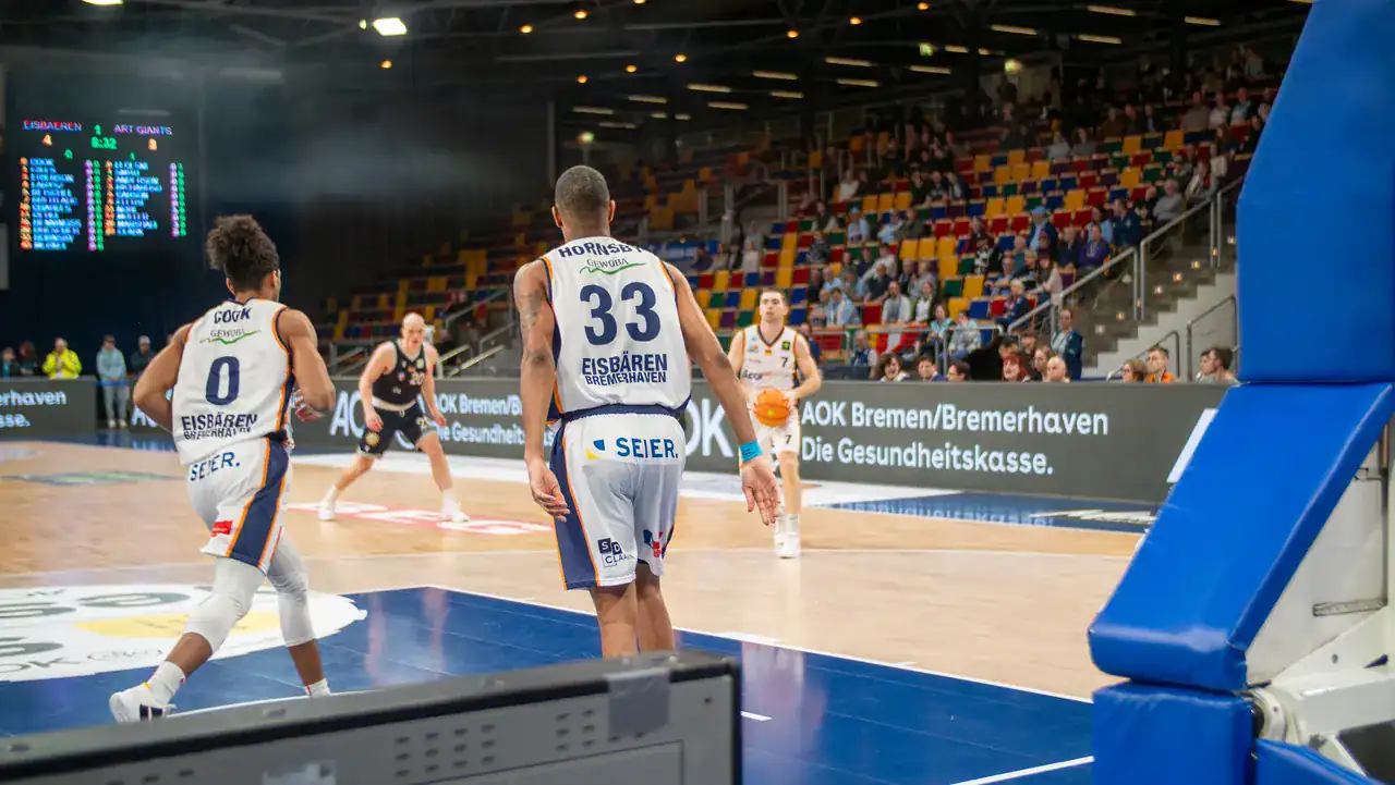 Blick hinter dem Korb auf Basketballspieler und die Zuschauertribüne.